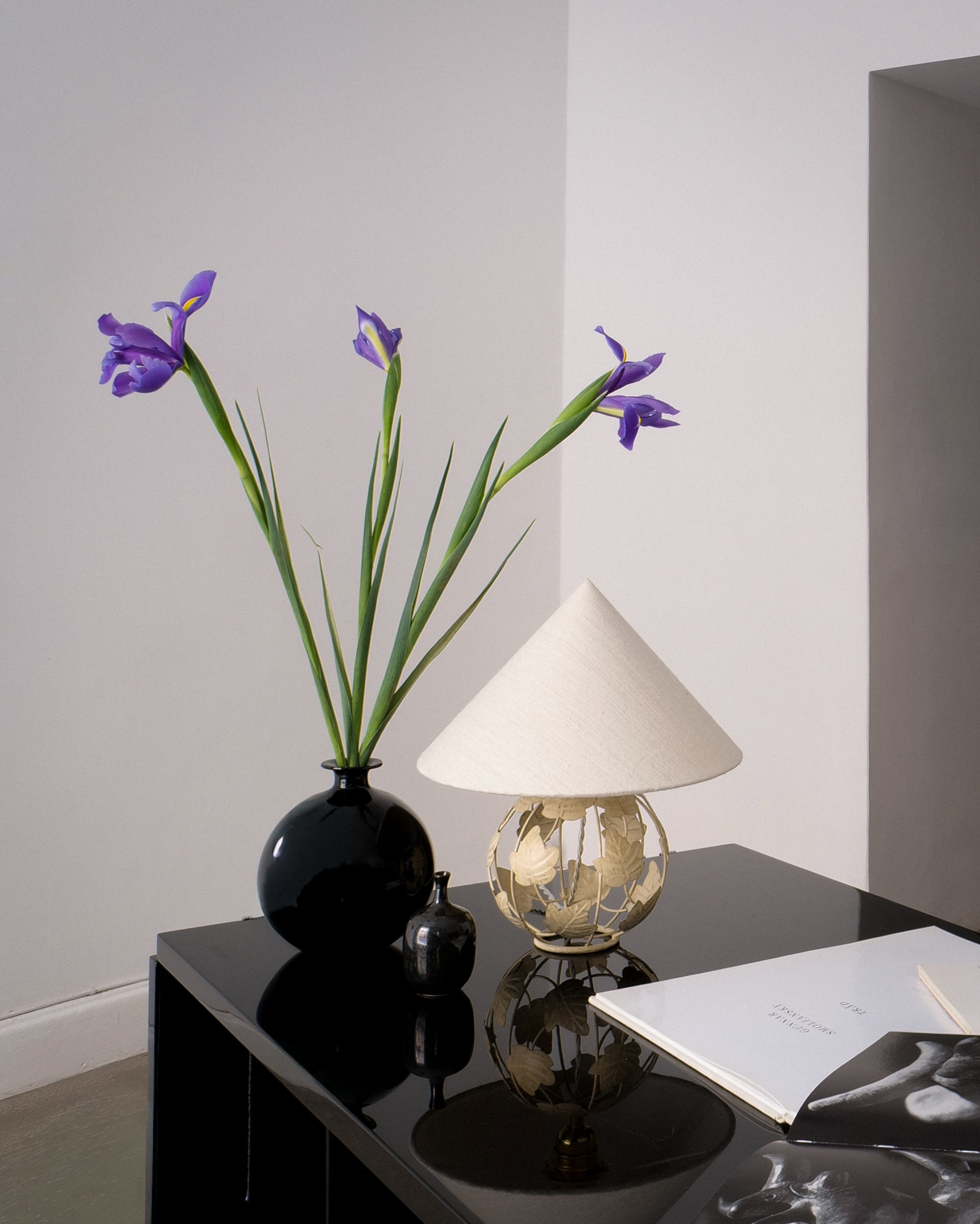Decorative table with a vase of purple flowers, a lamp, and a book against a plain wall.