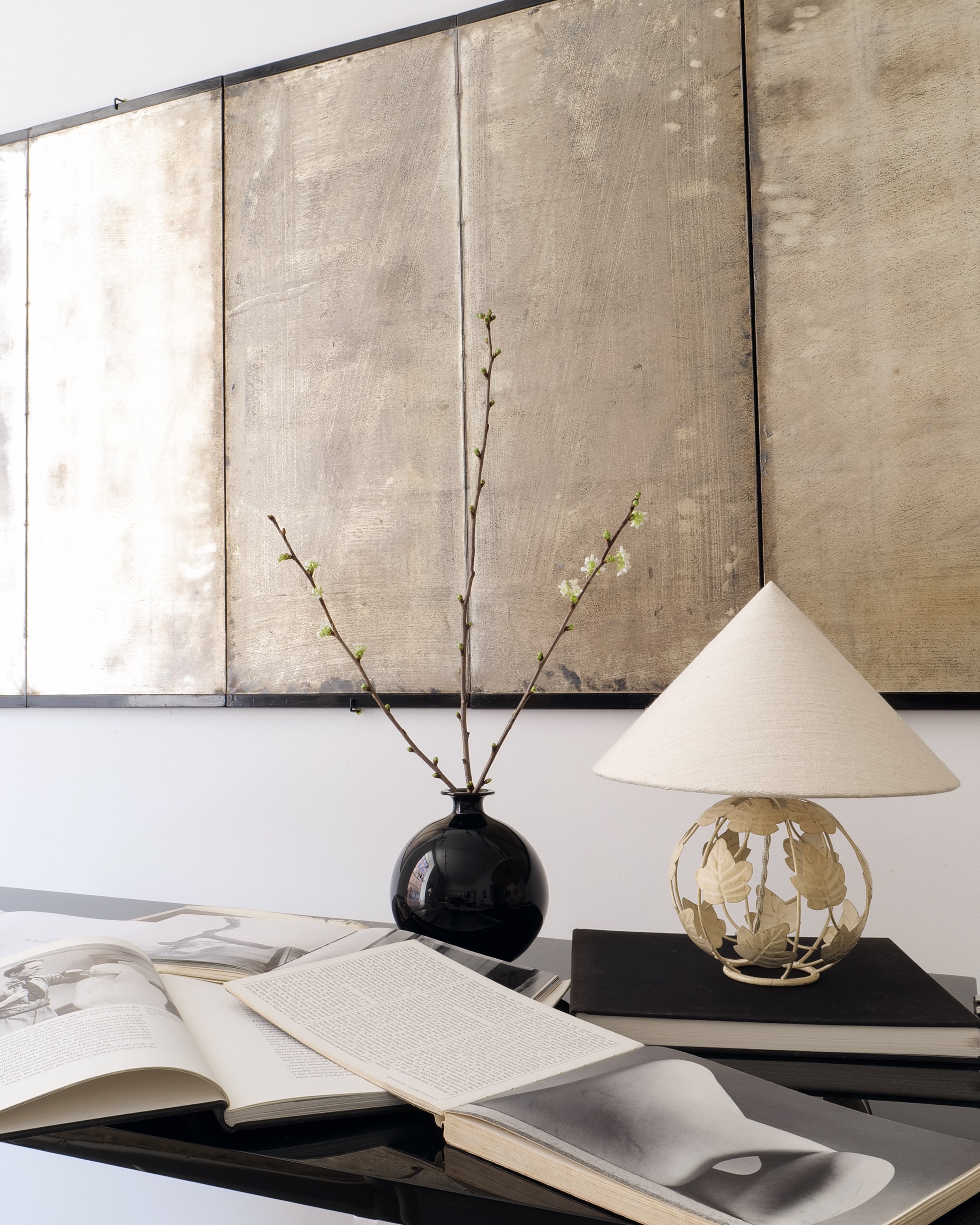Decorative setting with books, a vase with branches, and a lamp against a textured wall.