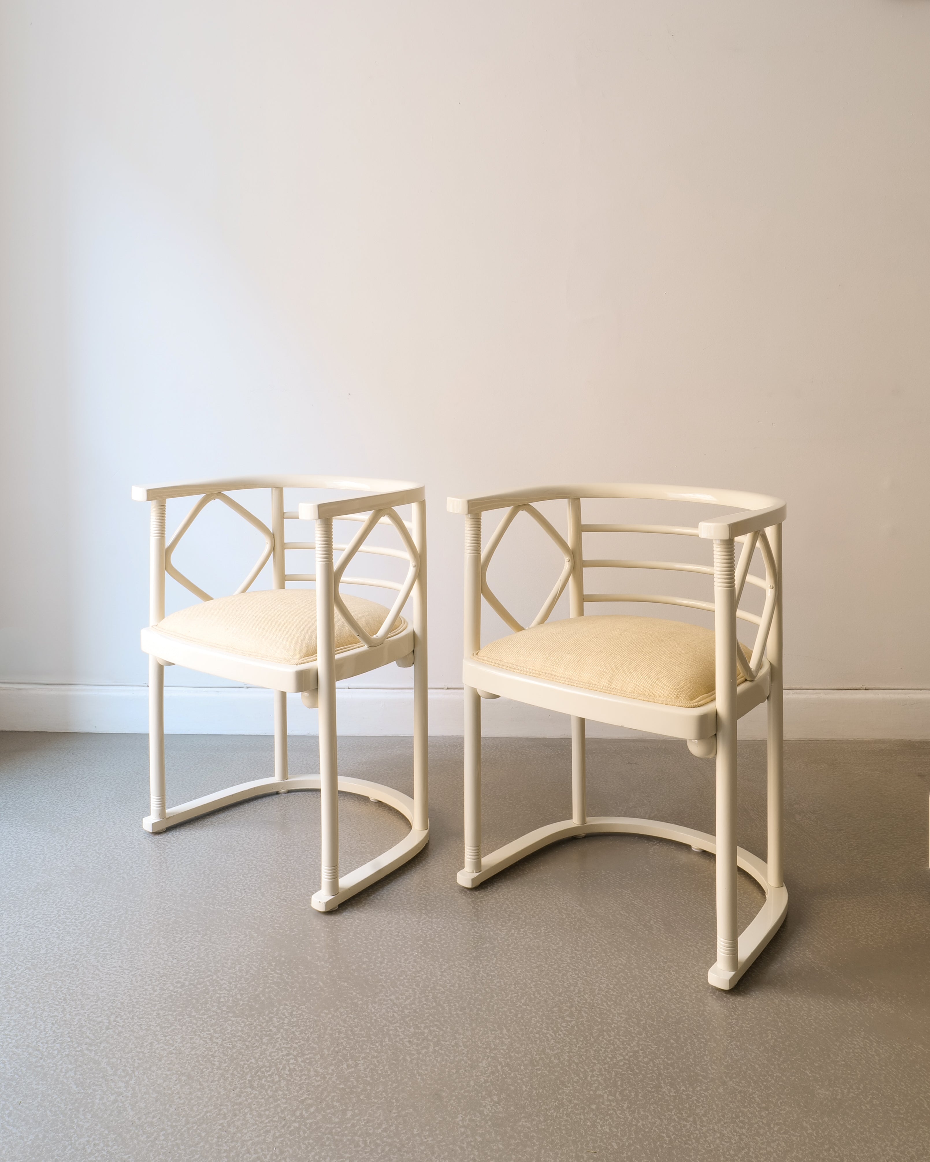 Two off white lacquered Josef Hoffman chairs with beige seat on a plain floor and wall background