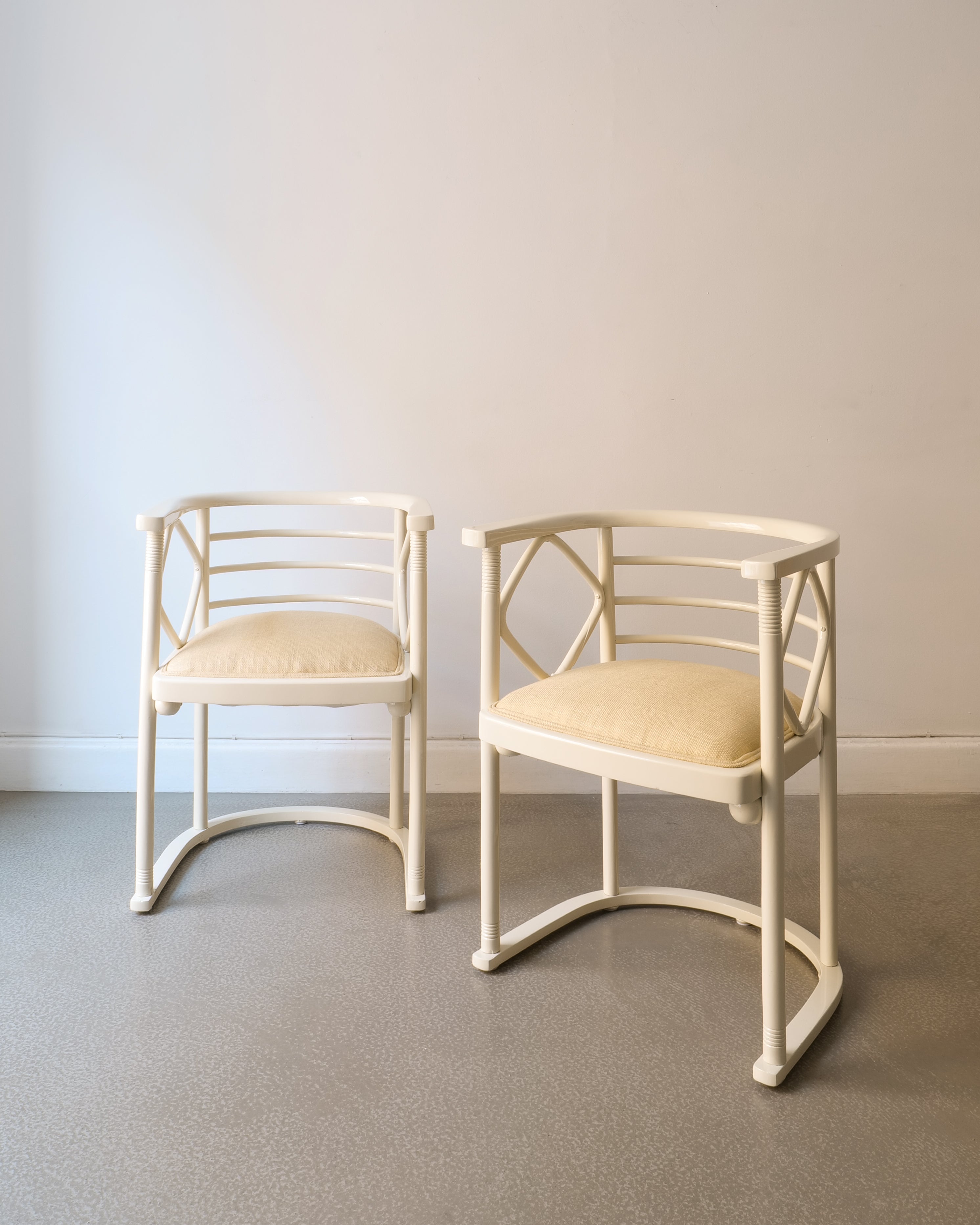 Two off white lacquered Josef Hoffman chairs with beige seat on a plain floor and wall background