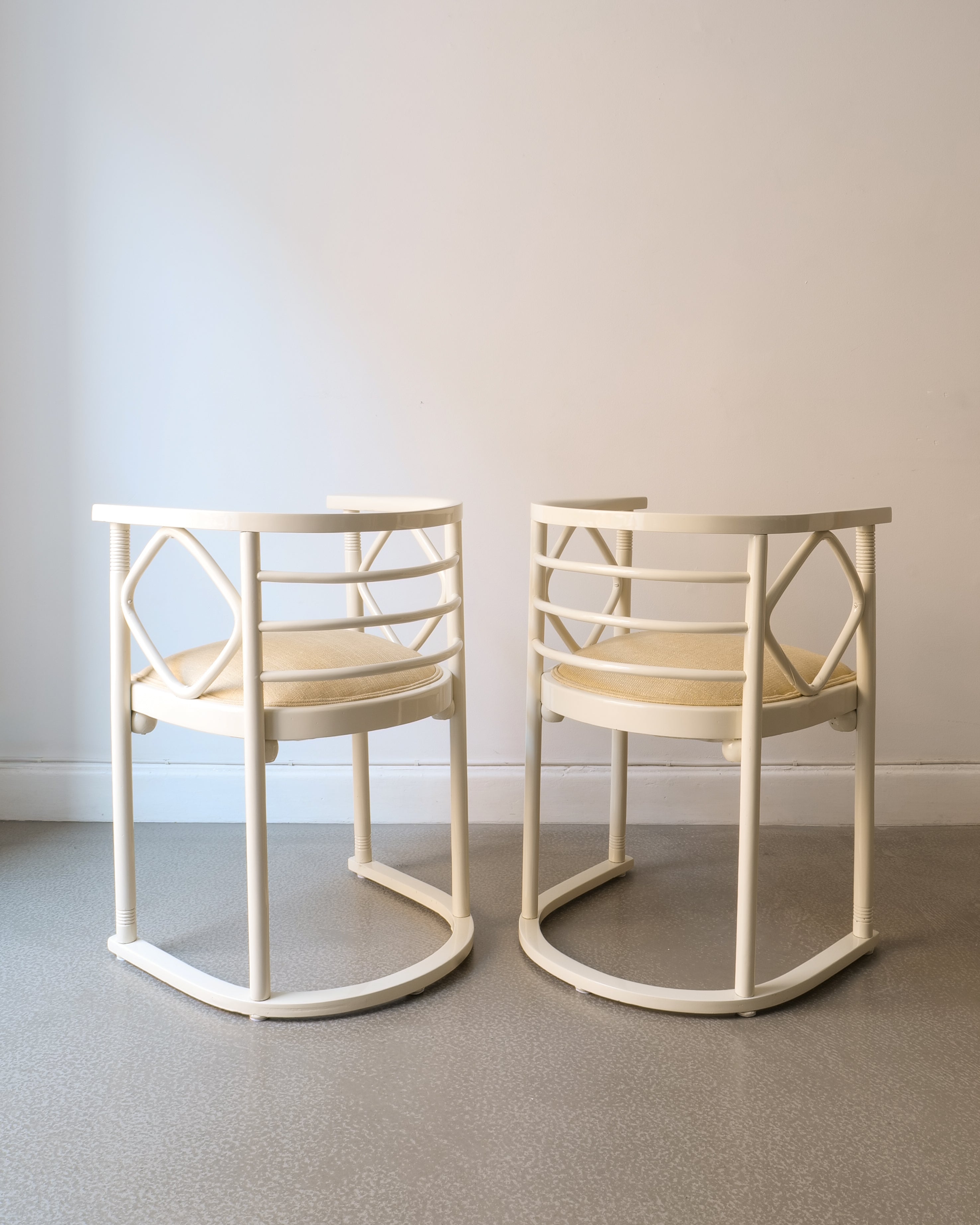 Two off white lacquered Josef Hoffman chairs with beige seat on a plain floor and wall background
