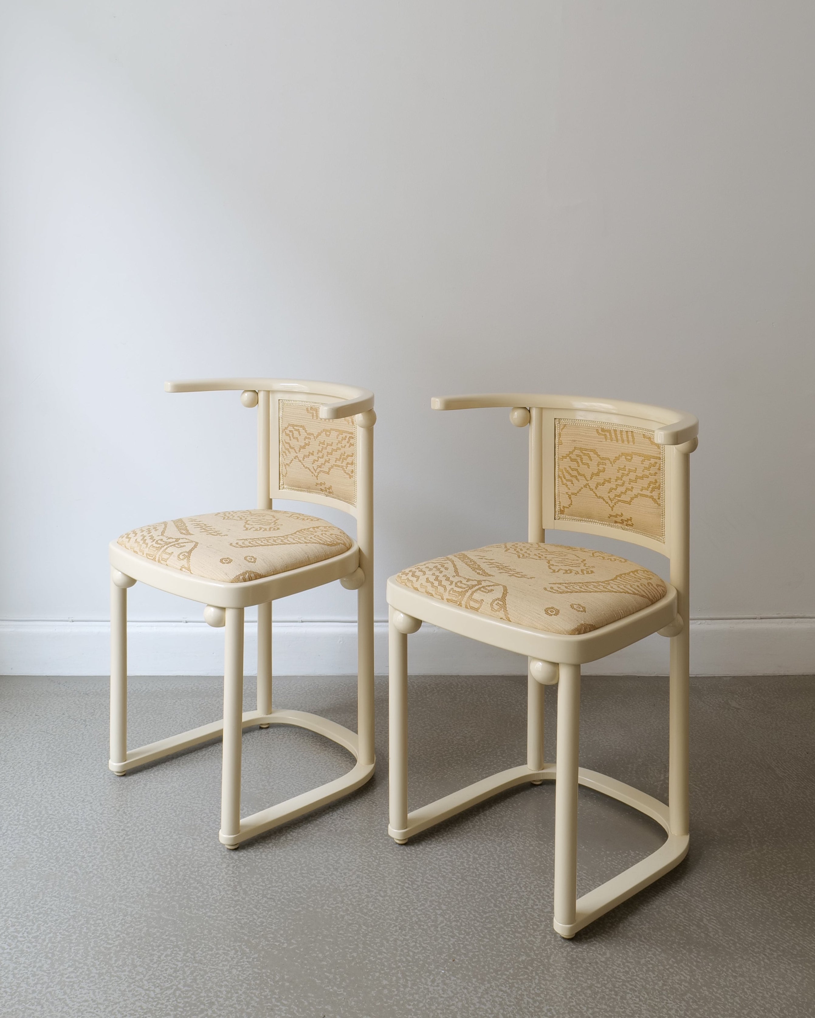 A pair of beige upholstered slipper chairs with a high backrest and intricate wood details, set against a neutral background.