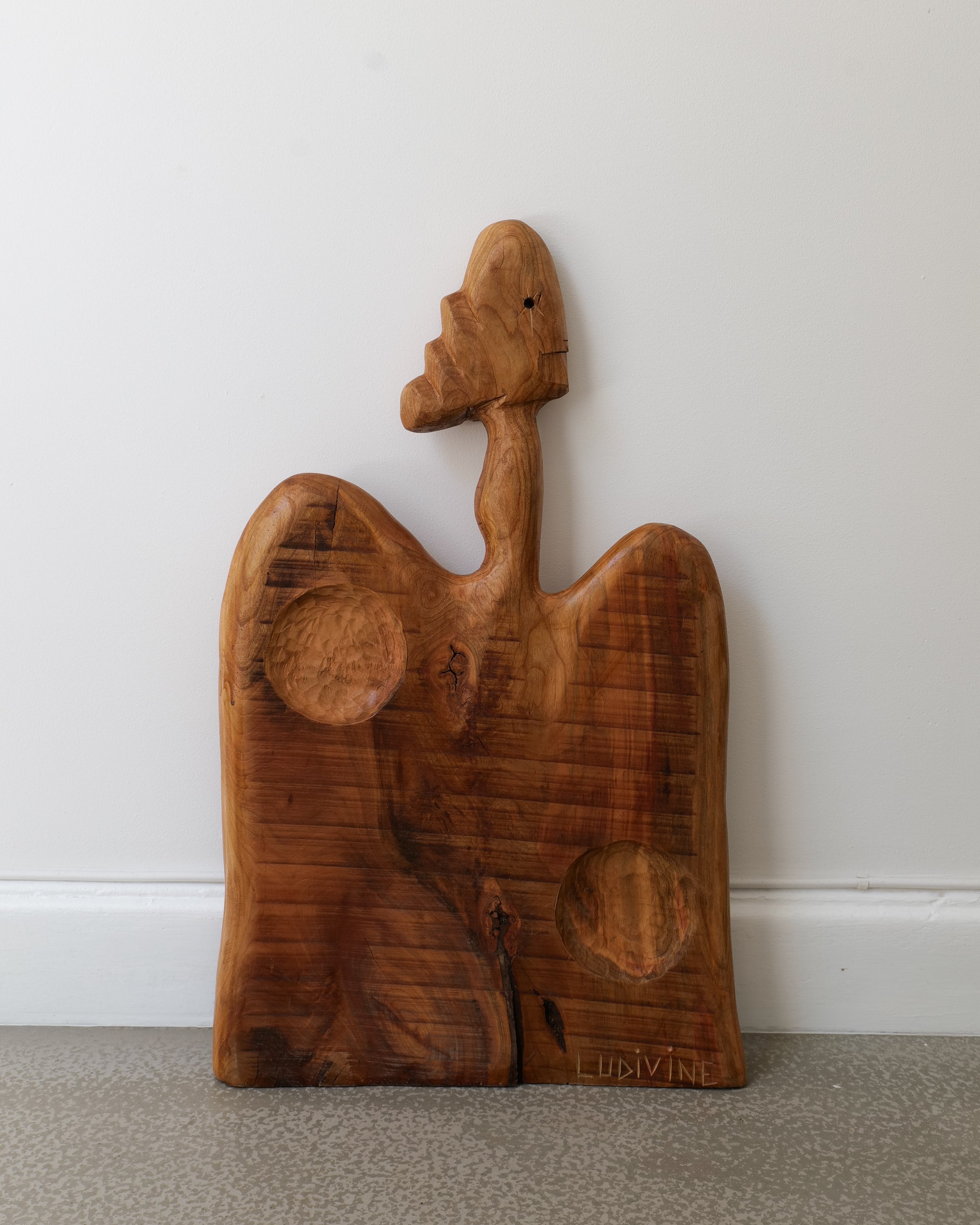 A wooden chopping board with a sculptural handle in the shape of a stylized animal or figure, handcrafted and posed as if cut out of a tree.