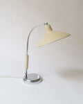 A 1930s functionalist desk lamp with a chrome and painted metal body in a buttery cream color, featuring a moveable arm and a bakelite lamp holder.