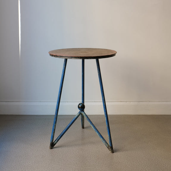 A vintage metal table with a tripod form and a round wooden top, featuring blue painted legs and a patina finish.