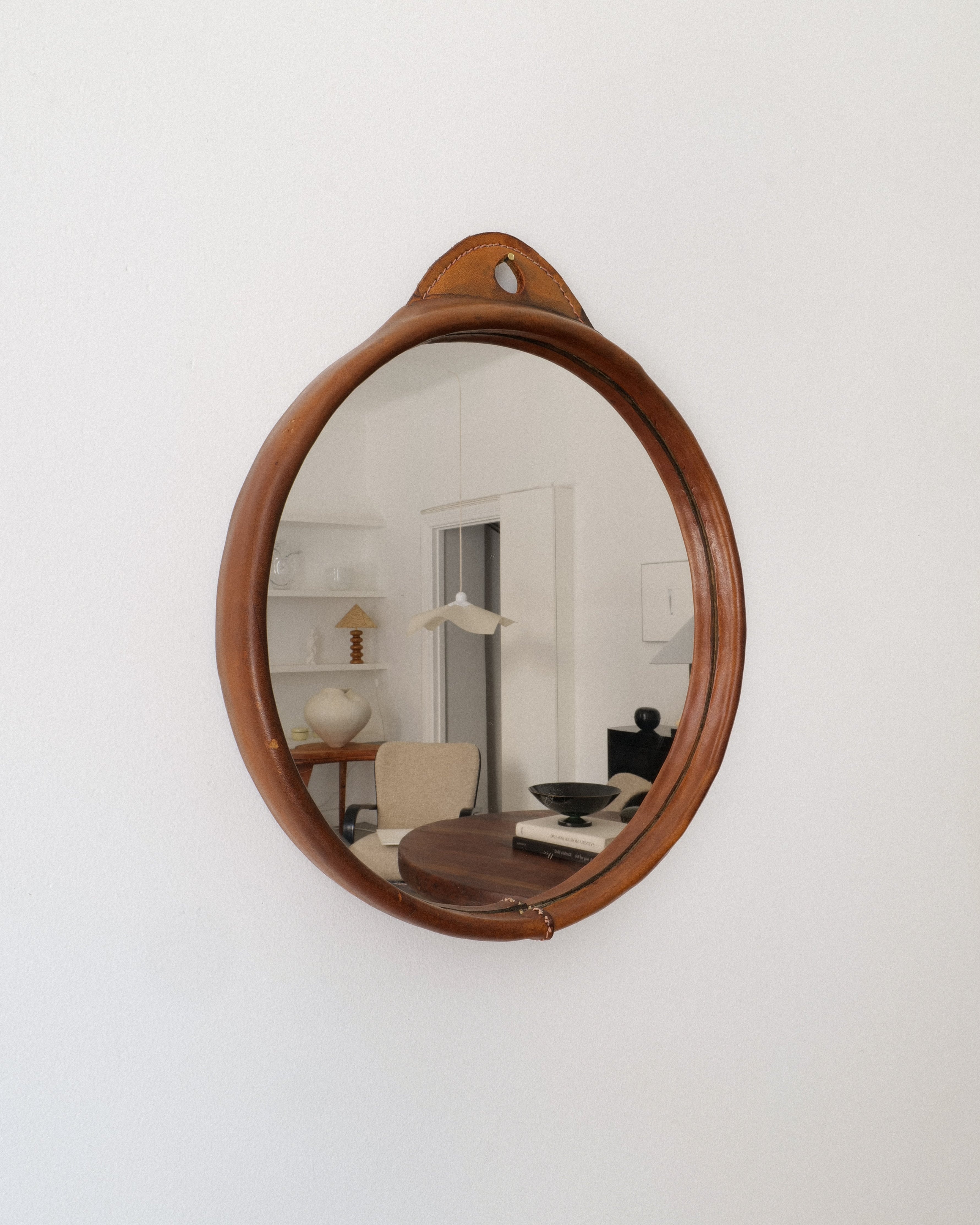 A circular mirror with a brown saddle-stitched leather frame, hanging on a wall.