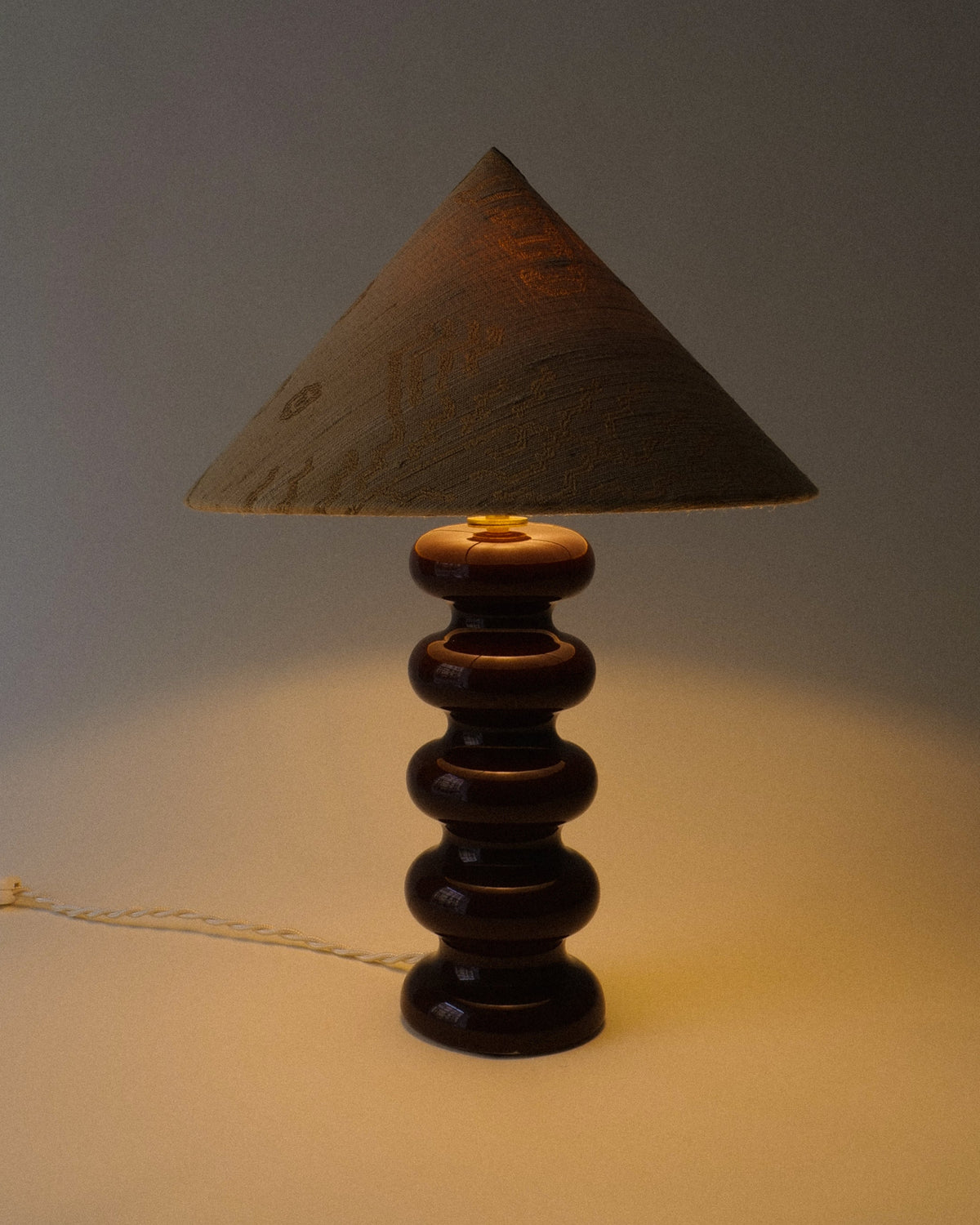 Doulton stacked sphere ceramic table lamp 1970s, bitter chocolate glaze with gold patterned conical shade, full view lit in warm setting.