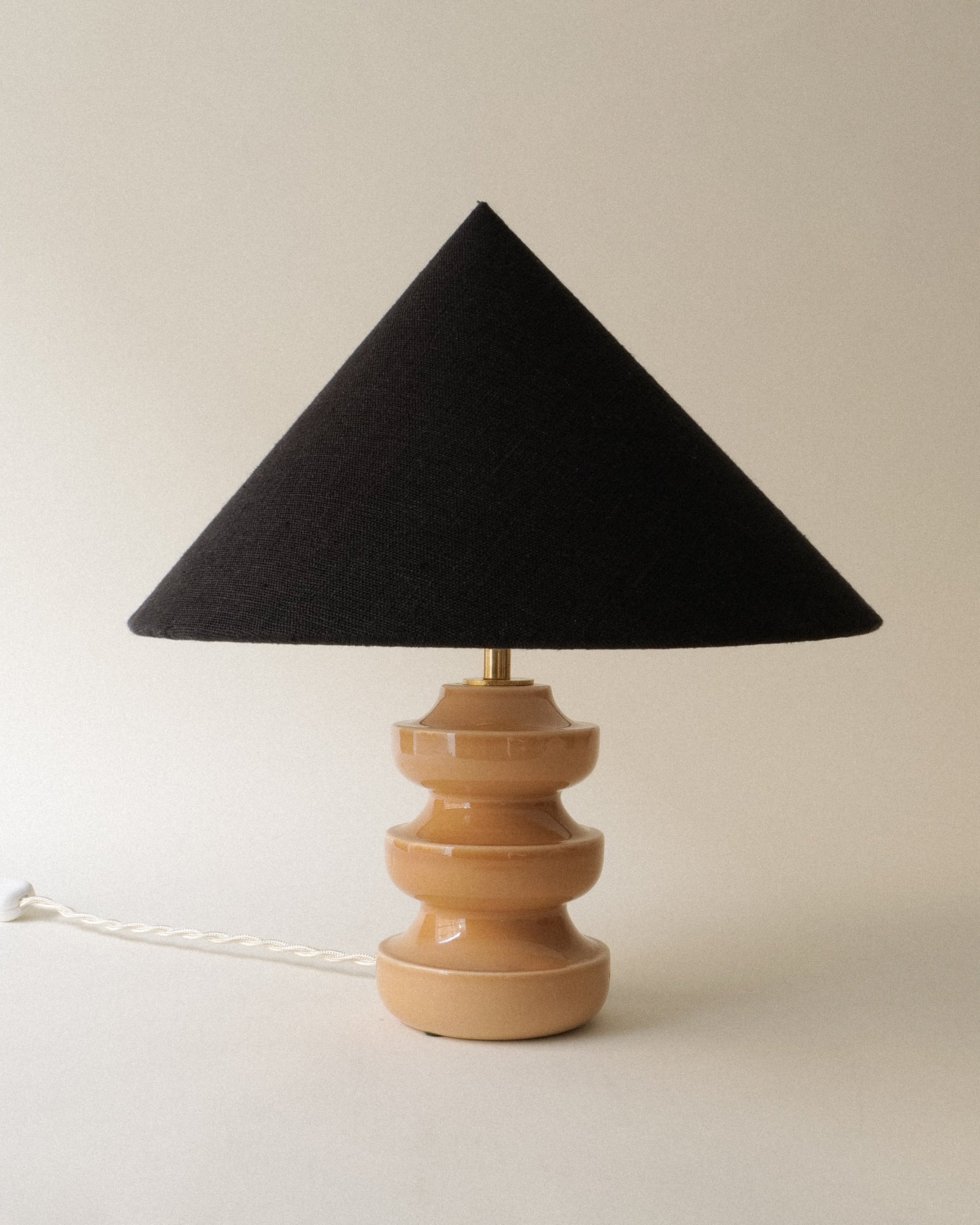 A ceramic table lamp with a light blush pink base and a black, pointy lampshade. The lamp is turned off, and the image is taken against a neutral background.