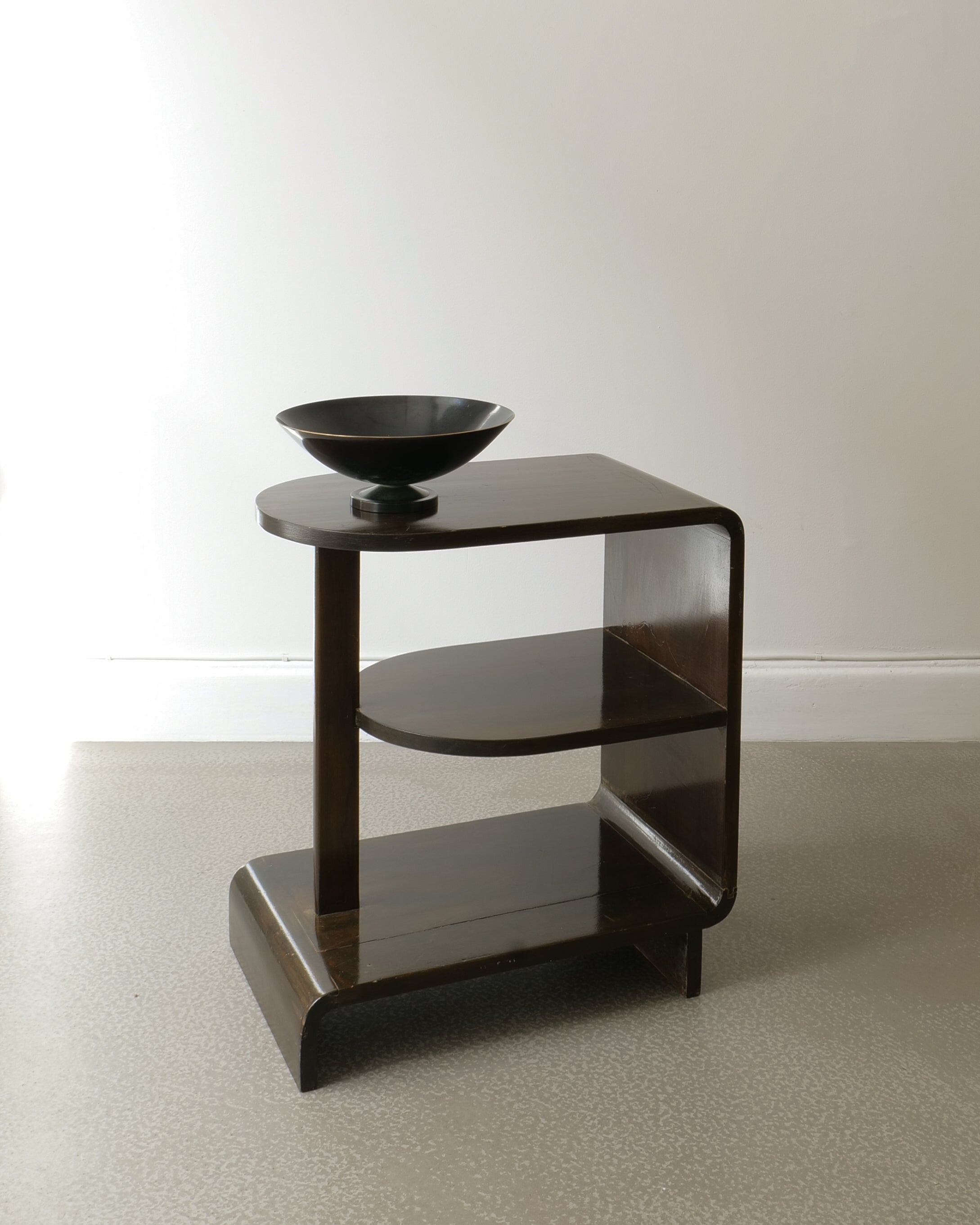 Tiered wood side table with curved edges with metal bowl and plain background.