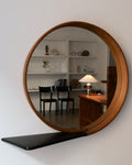 A vintage Swedish Modern style circular mirror with brass studs and a shelf in stained black, reflecting a room with furniture in the background.