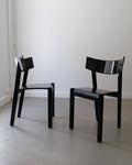 Two black lacquered wood chairs with a sleek design, featuring a high backrest, placed against a white wall on a light grey floor.