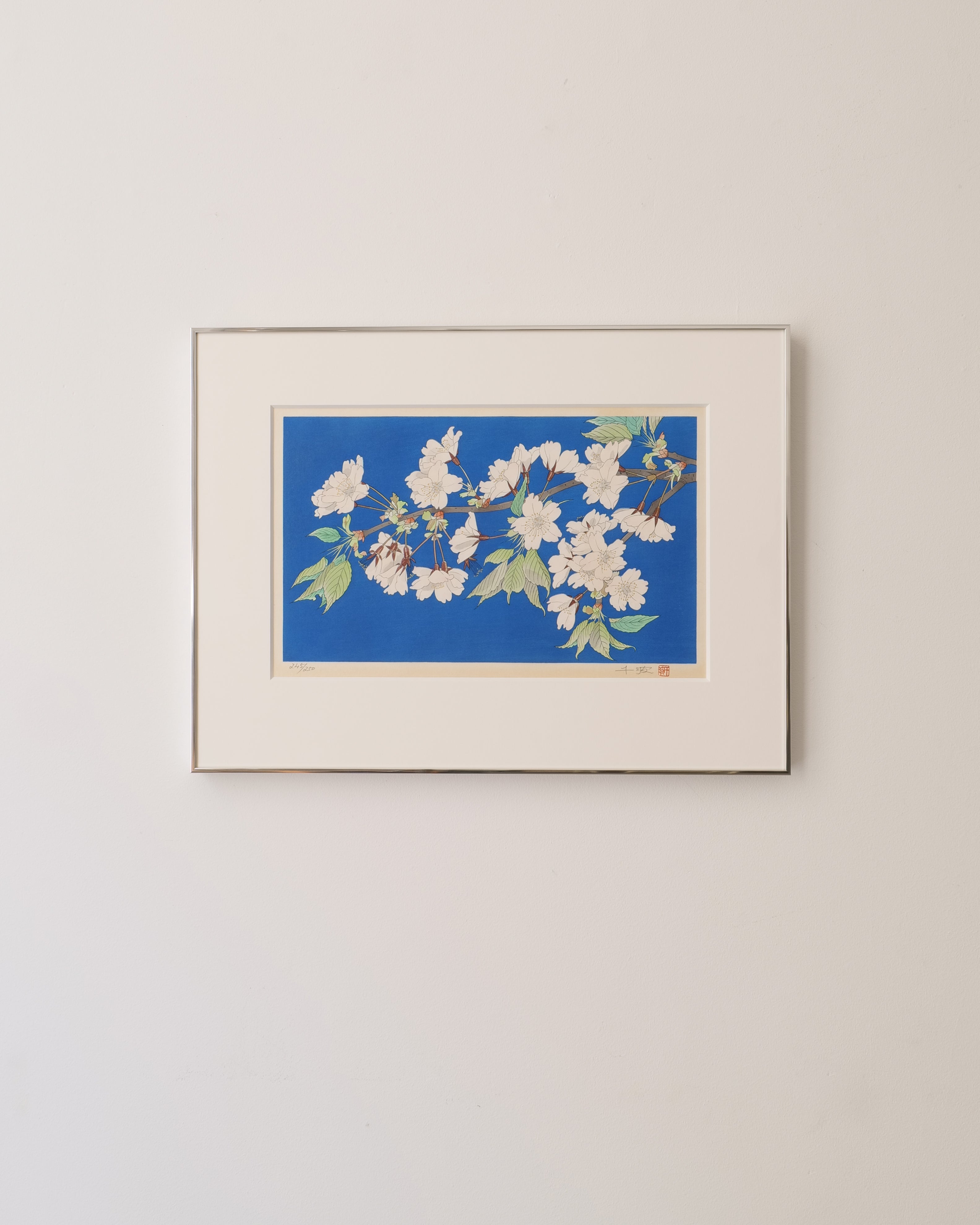Framed Japanese wood block printed artwork of a floral blossom design on a blue background, mounted on a beige wall.