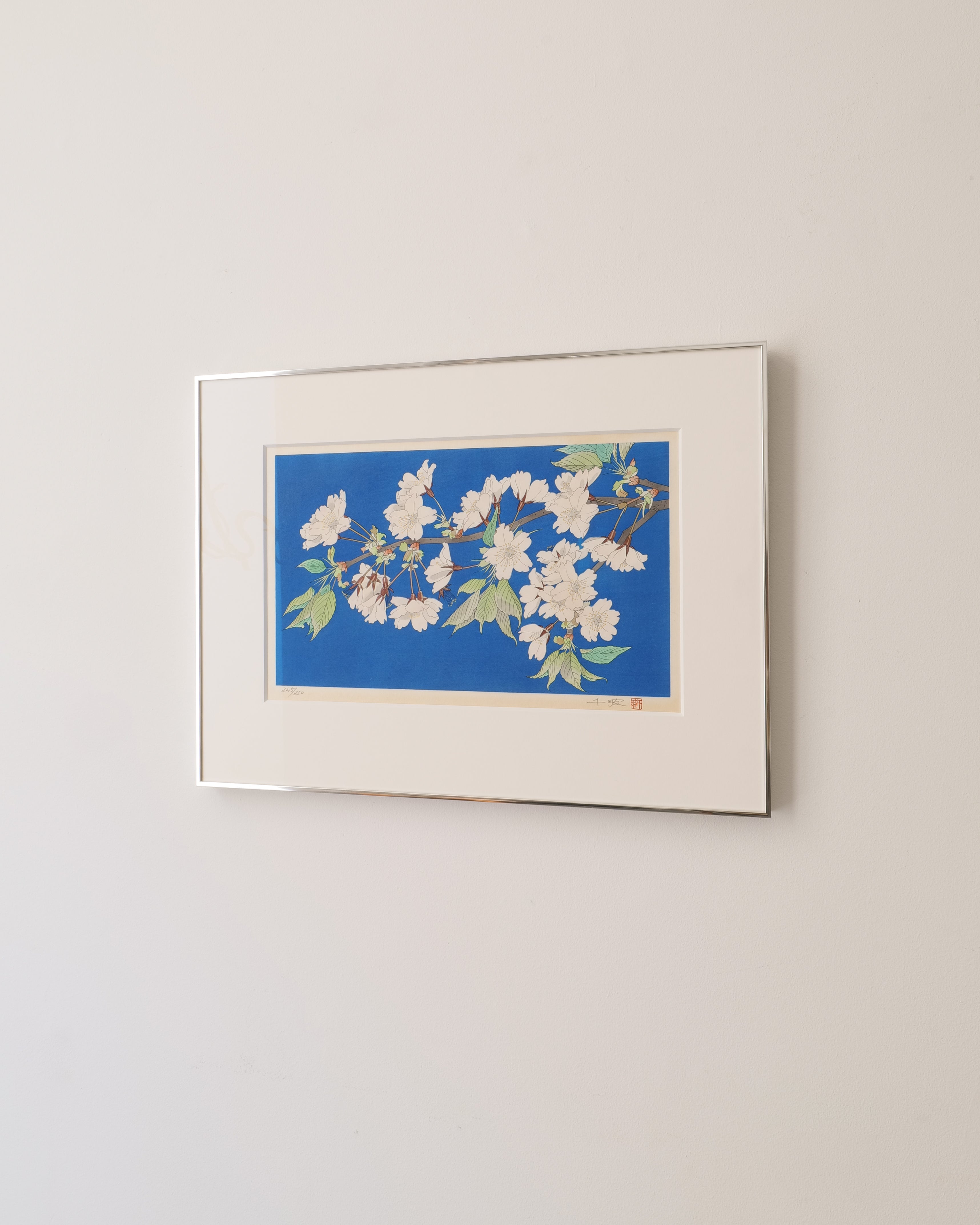 Framed Japanese wood block printed artwork of a floral blossom design on a blue background, mounted on a beige wall.