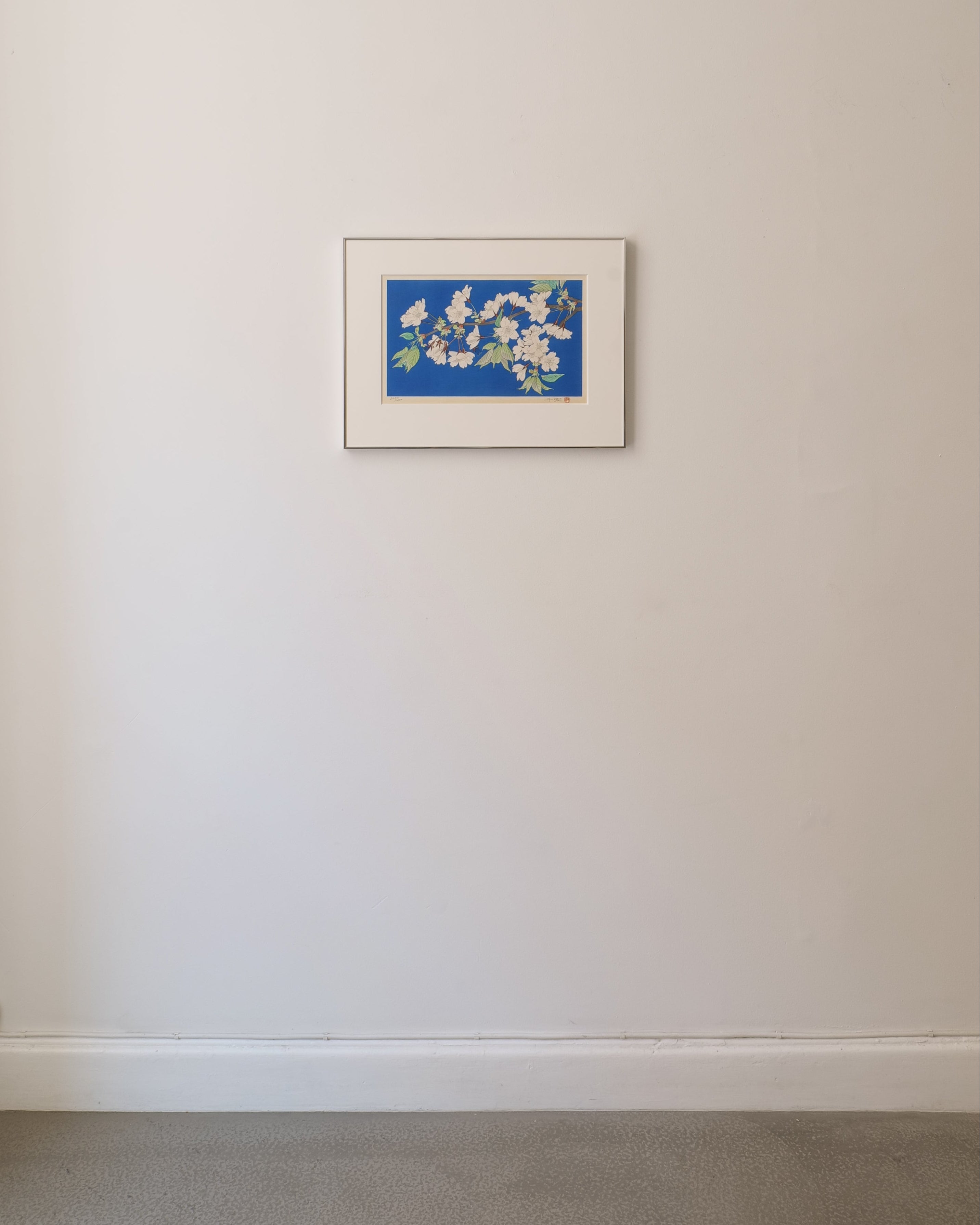 Framed Japanese wood block printed artwork of a floral blossom design on a blue background, mounted on a beige wall.